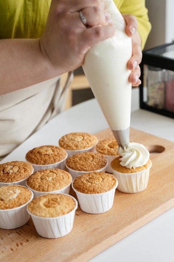 High angle of crop unrecognizable confectioner with pastry bag squeezing white cream on cupcakes