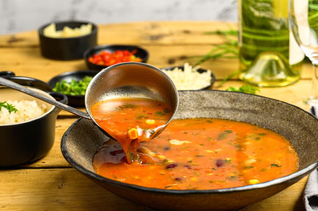 Gros plan d'une louche servant de la soupe aux légumes dans un bol rustique avec du vin et des plats d'accompagnement.