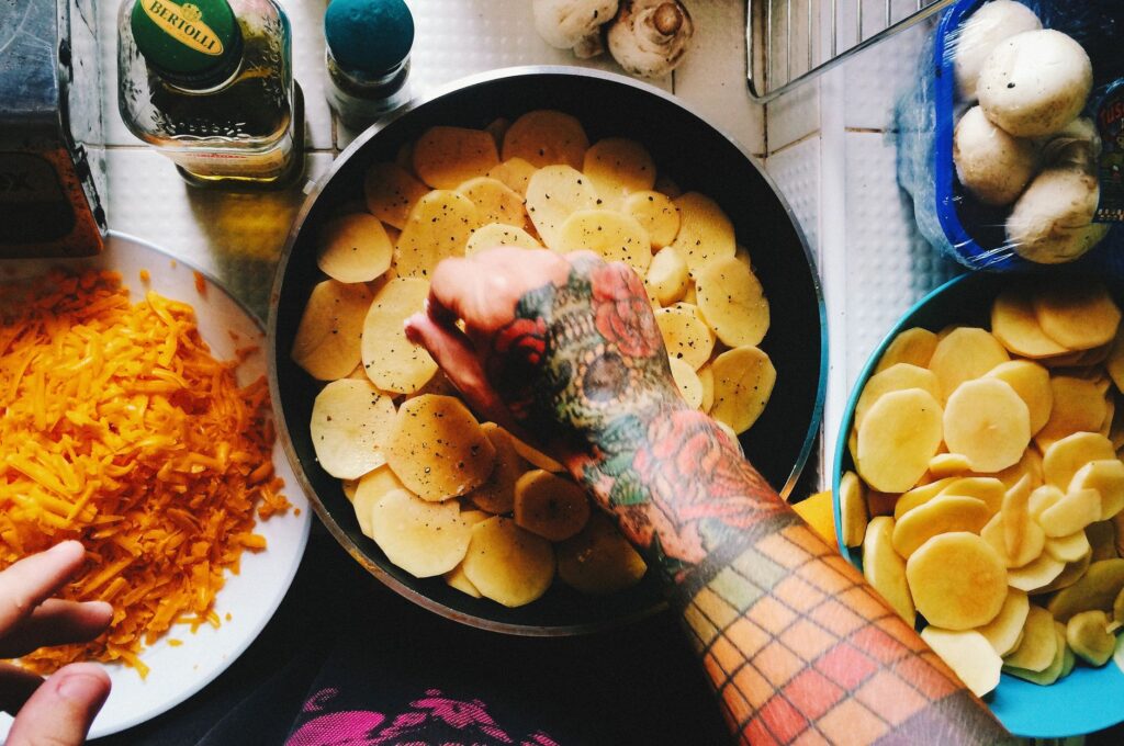 Vue en plongée d'un bras tatoué préparant un plat de pommes de terre et de fromage sur un plan de travail de cuisine.