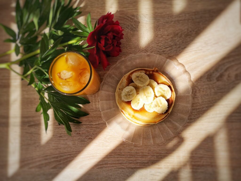 Vue de dessus de pancakes à la banane et de jus d'orange avec une fleur rouge, créant une ambiance de petit déjeuner chaleureuse.