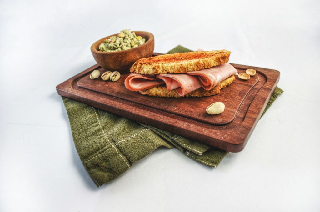 Sandwich gourmet au jambon avec des pistaches et une sauce sur une planche en bois, parfait pour la photographie culinaire.
