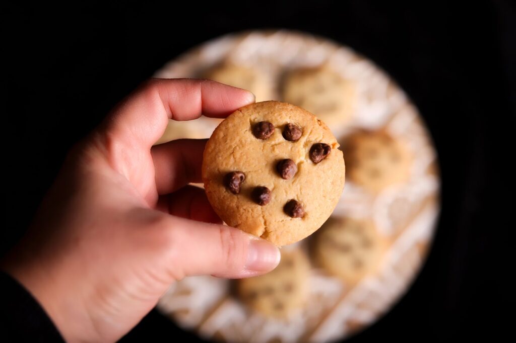 Gros plan sur une main tenant un cookie aux pépites de chocolat au-dessus d'une assiette floue de cookies sur un fond sombre.