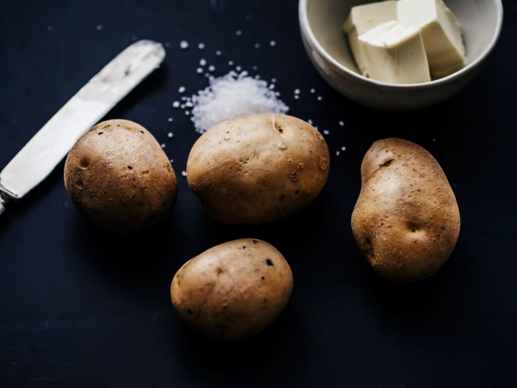 Nature morte artistique présentant des pommes de terre, du beurre et du sel sur un fond sombre.