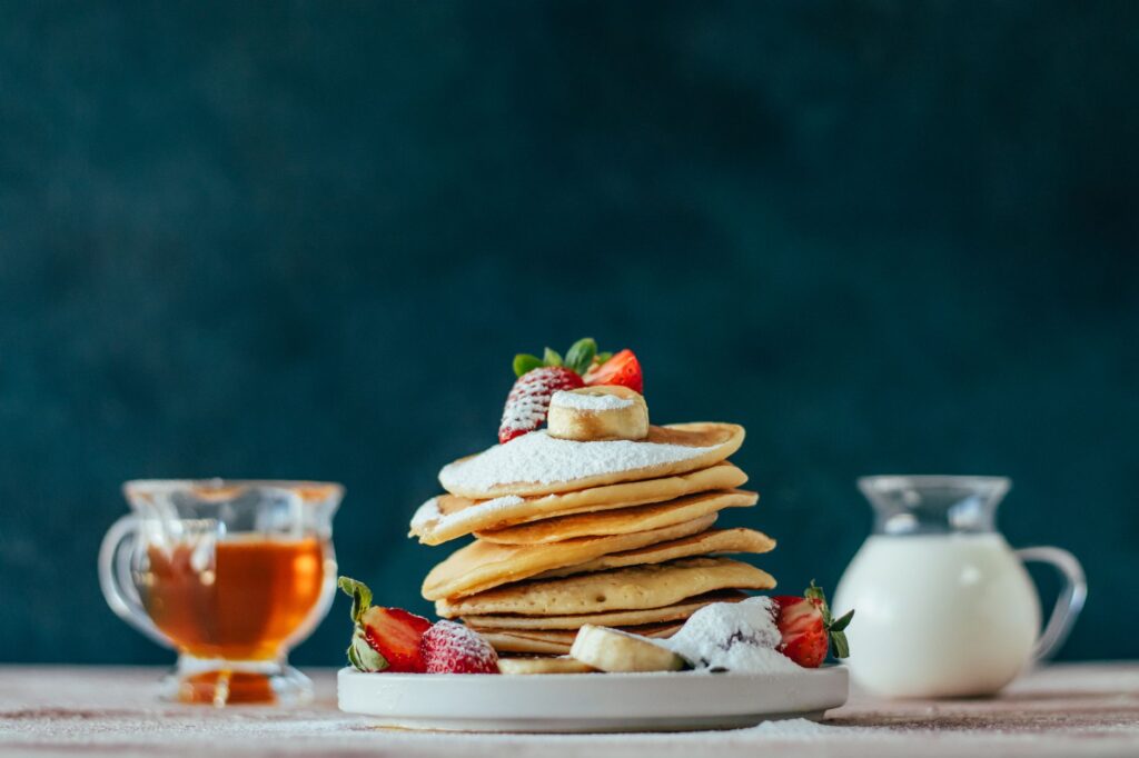 Une pile de crêpes moelleuses garnies de fruits, de sucre en poudre, de miel et de crème.