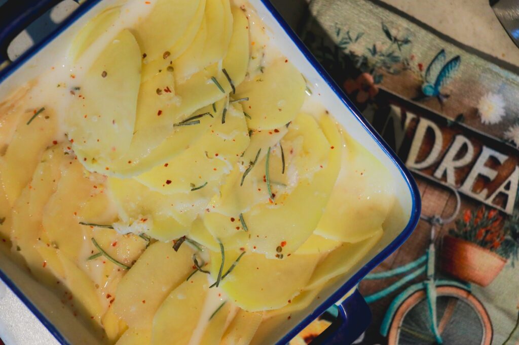 Gros plan sur des pommes de terre finement tranchées, assaisonnées d'herbes et d'épices, présentées dans un plat en émail bleu, prêtes à être cuites au four.
