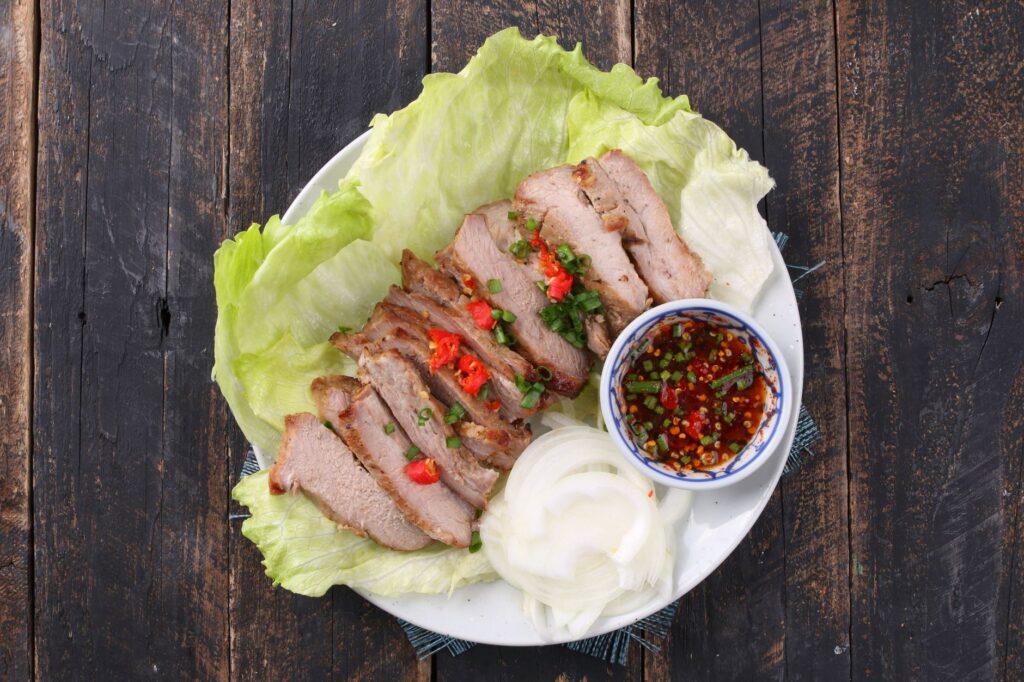 Assiette de porc grillé en tranches sur un lit de laitue, avec oignons et sauce piquante, présentée sur une table rustique en bois.