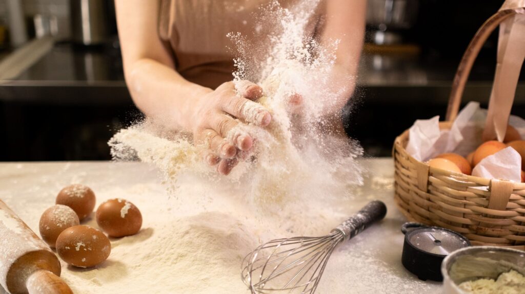 Dans une cuisine chaleureuse, un boulanger pétrit de la pâte entouré d'œufs, de farine et d'ustensiles de cuisine.