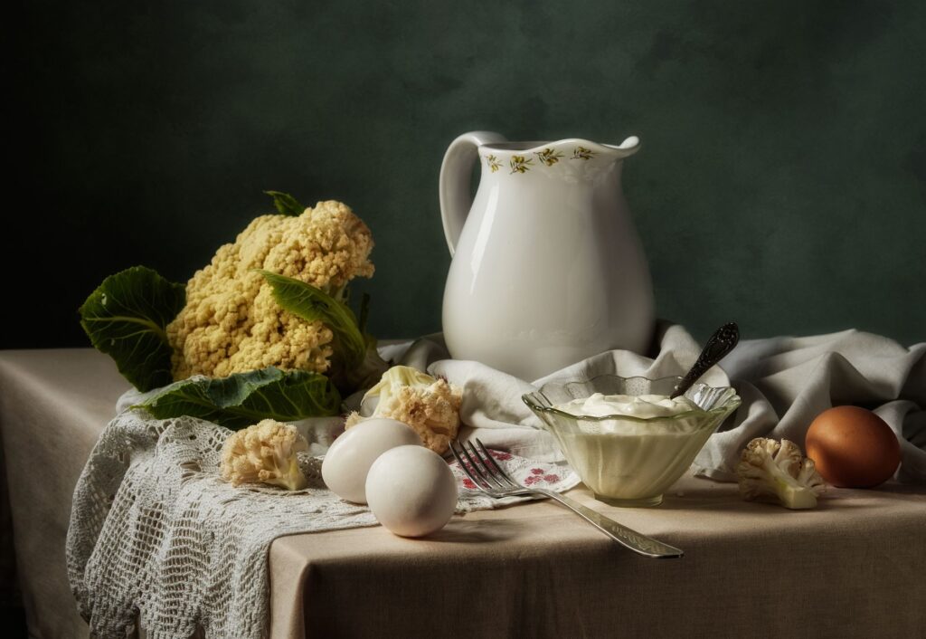 Une nature morte classique représentant un chou-fleur, des œufs et un pichet dans un décor de cuisine.