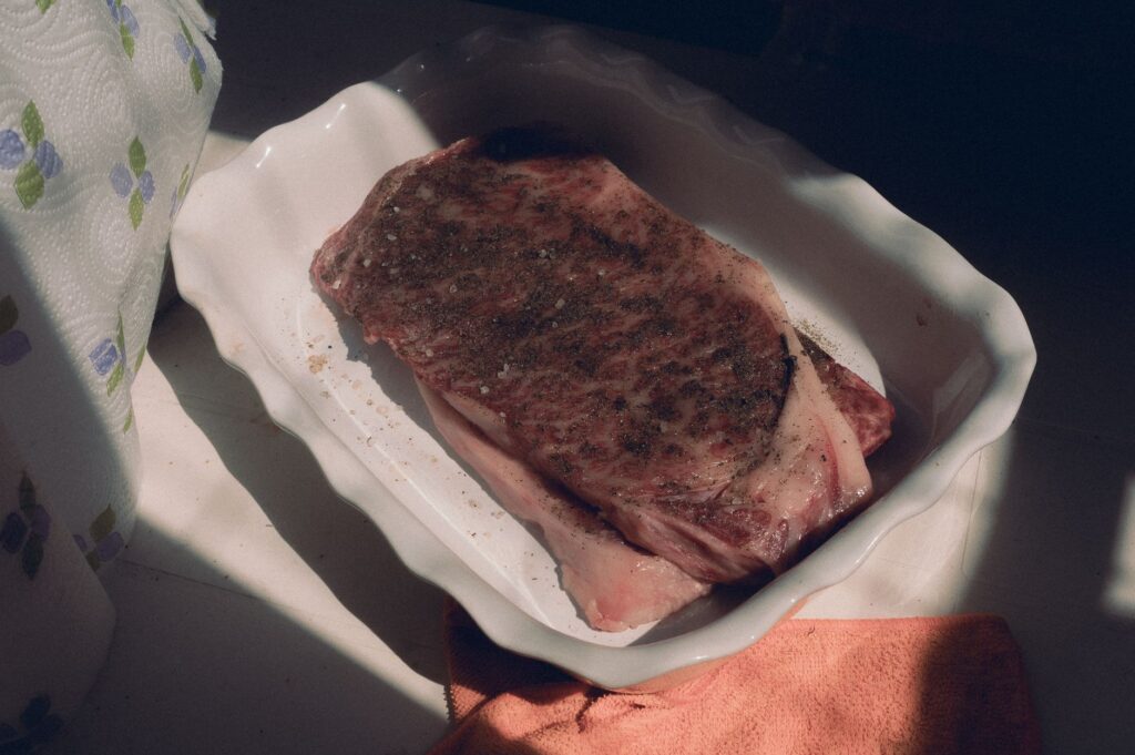Un steak de bœuf cru persillé, assaisonné et prêt à cuire dans un plat allant au four, baigné de lumière naturelle.