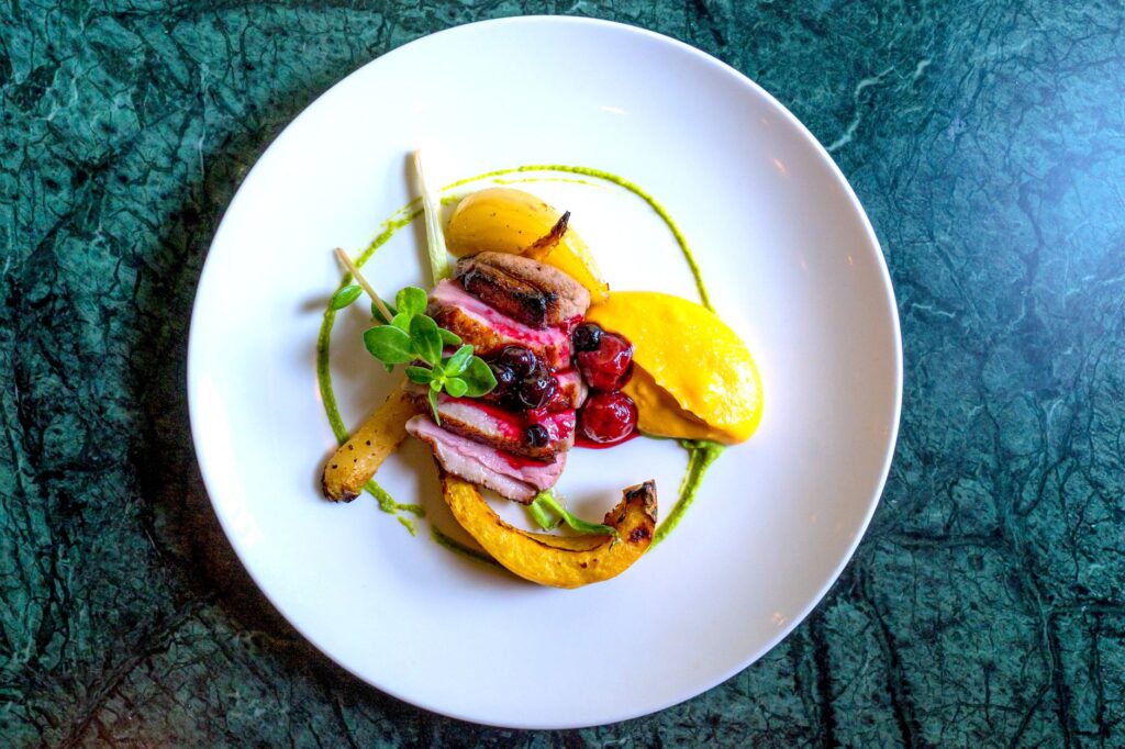 Présentation artistique d'un magret de canard avec sauce aux baies, légumes racines et purée sur une assiette blanche.