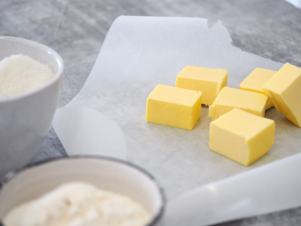 Gros plan de cubes de beurre sur du papier sulfurisé avec des bols d'ingrédients dans une cuisine.
