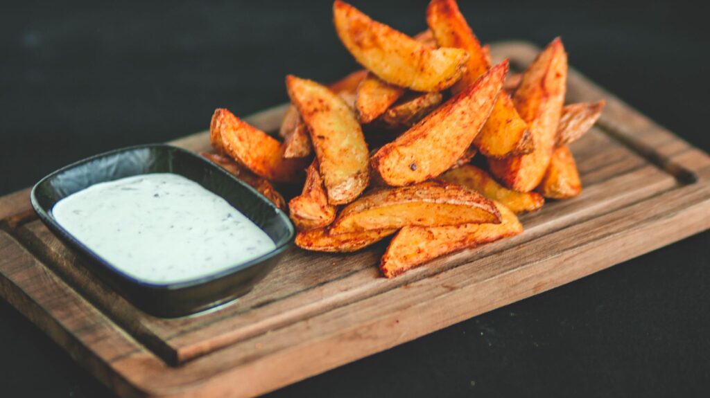 Des quartiers de pommes de terre frites dorées servis avec une sauce crémeuse sur une planche en bois.