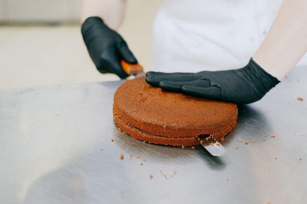 Un pâtissier portant des gants tranche habilement un gâteau au chocolat avec une spatule sur une table en métal.