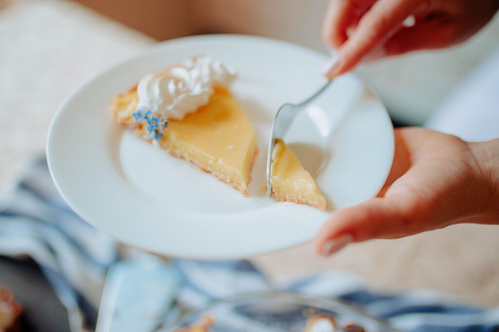 Un gros plan d'une main servant une part de tarte au citron maison avec de la crème fouettée sur une assiette blanche.