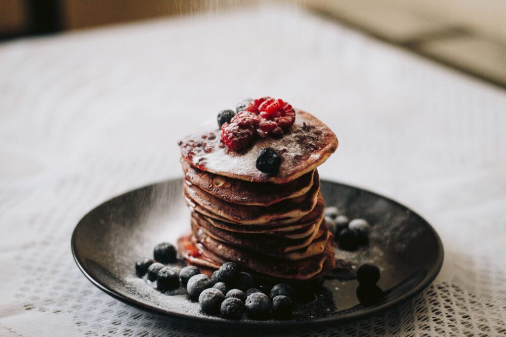 Empilement de pancakes moelleux ornés de bleuets frais et de framboises, saupoudrés de sucre.
