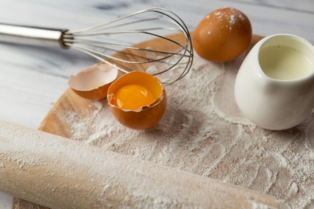 Un gros plan des essentiels de la pâtisserie comprenant des œufs, de la farine et un fouet sur une planche en bois.