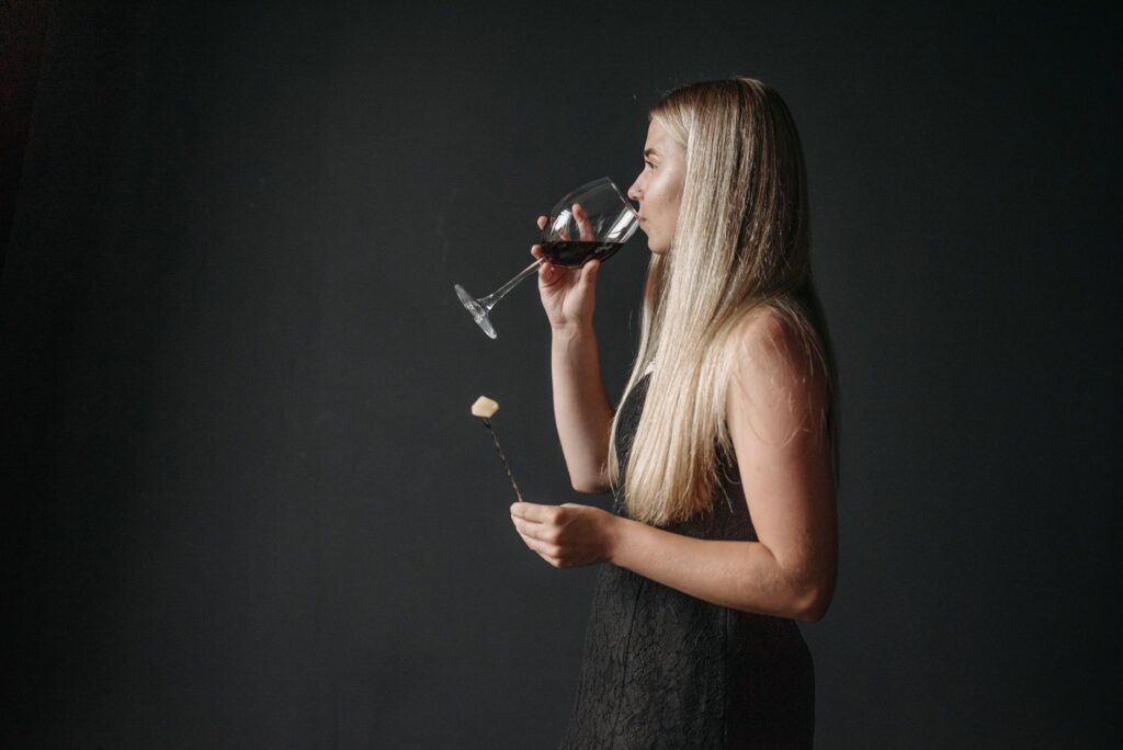 Femme blonde savourant du vin rouge tout en tenant une fourchette à fondue au fromage dans un cadre intérieur classique.