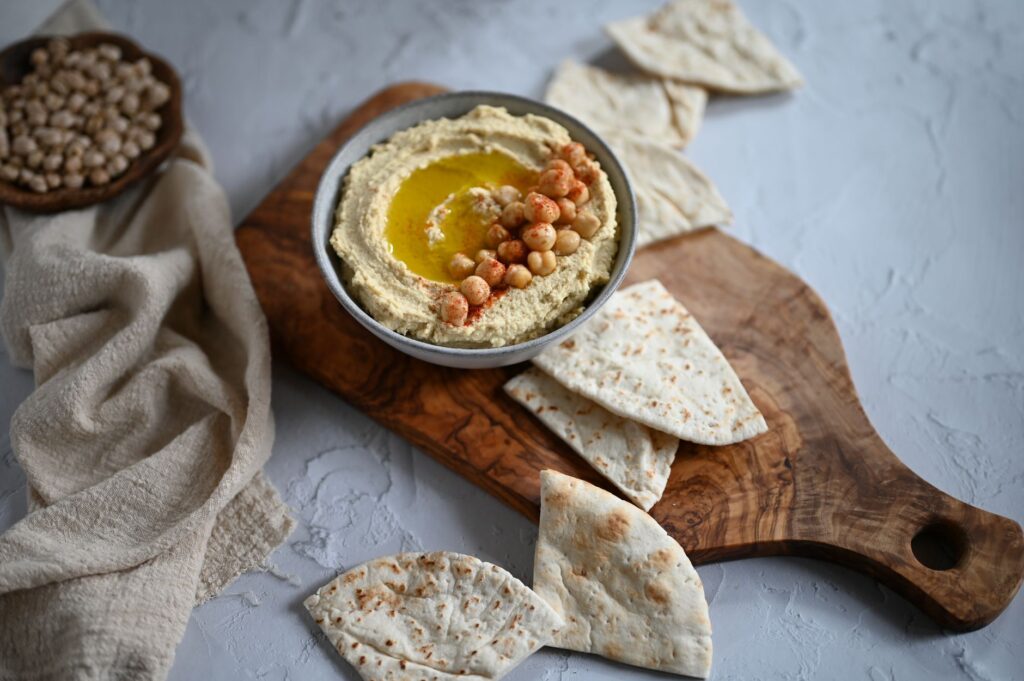 Un bol de houmous crémeux garni de pois chiches et d'huile d'olive, servi avec du pain pita sur une planche à découper en bois.