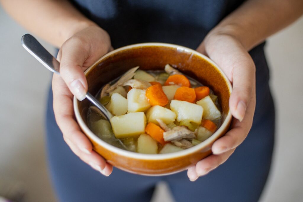 Un bol chaud de soupe de légumes maison tenu par des mains, mettant en valeur des carottes, des pommes de terre et du poulet.