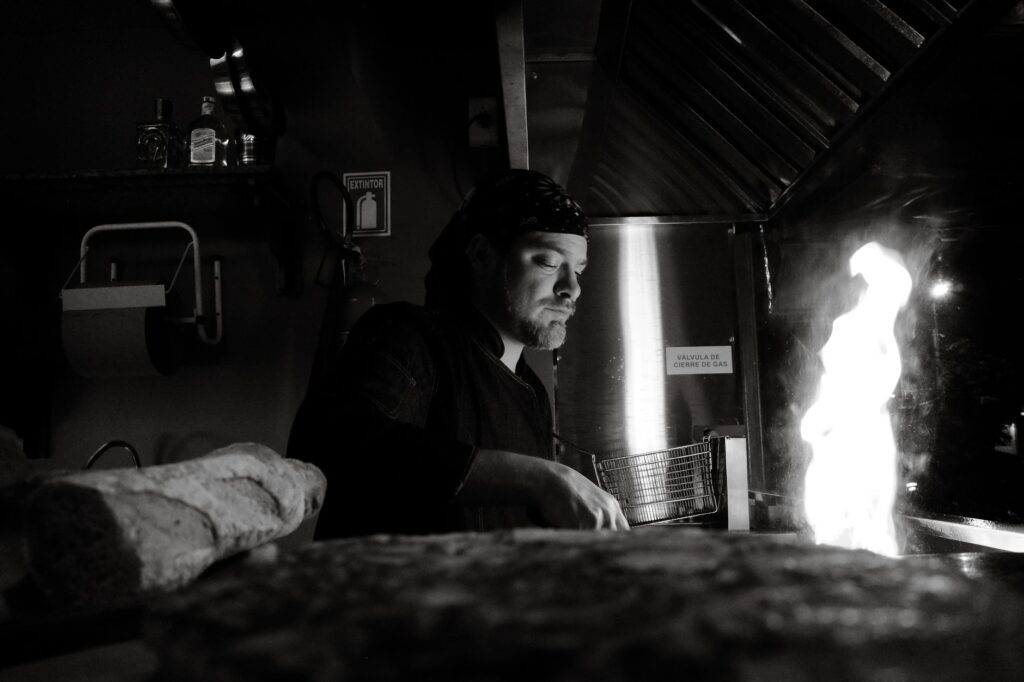 Un chef prépare habilement des plats au-dessus d'une flamme ouverte dans une cuisine commerciale, en noir et blanc.