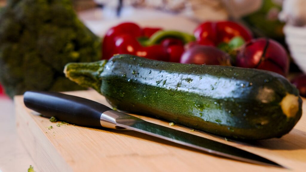 Gros plan sur une courgette posée sur une planche à découper, avec un couteau, entourée de légumes colorés.