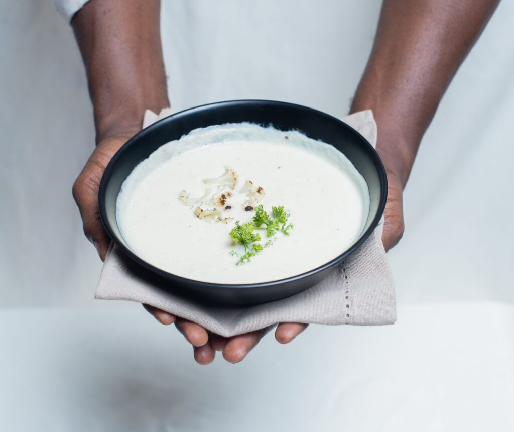 Un gros plan de mains tenant un bol de soupe crémeuse au chou-fleur garnie de persil.