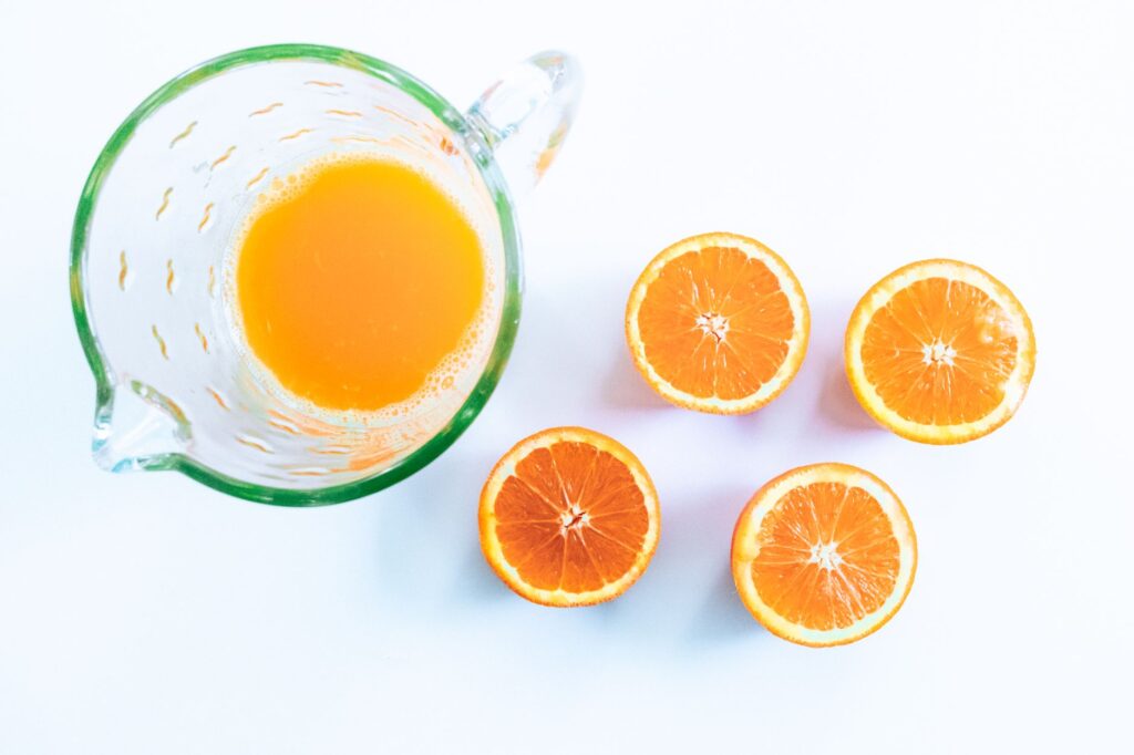 Carafe en verre de jus d'orange frais avec quatre oranges coupées en deux sur une surface blanche.