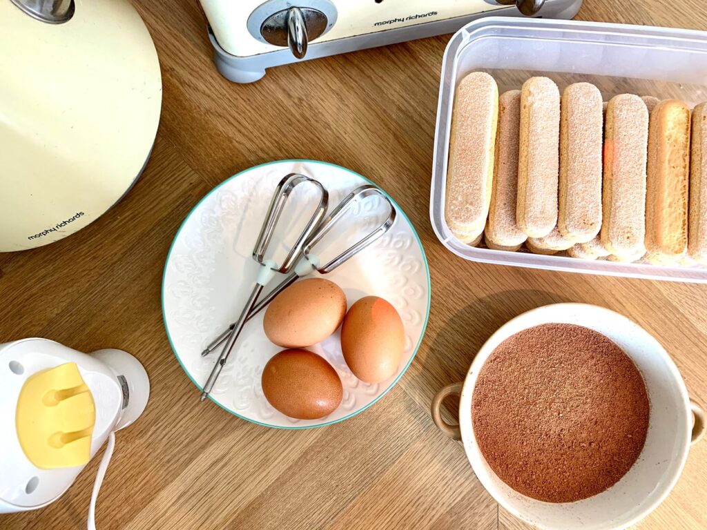 Une vue plongeante des ingrédients du tiramisu : œufs, cacao et biscuits à la cuillère sur une surface en bois.