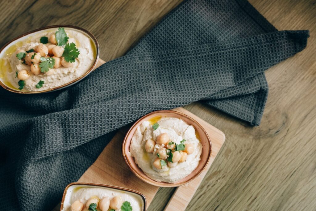 Vue de dessus de houmous crémeux fait maison dans des bols avec des pois chiches et des herbes sur une table en bois.
